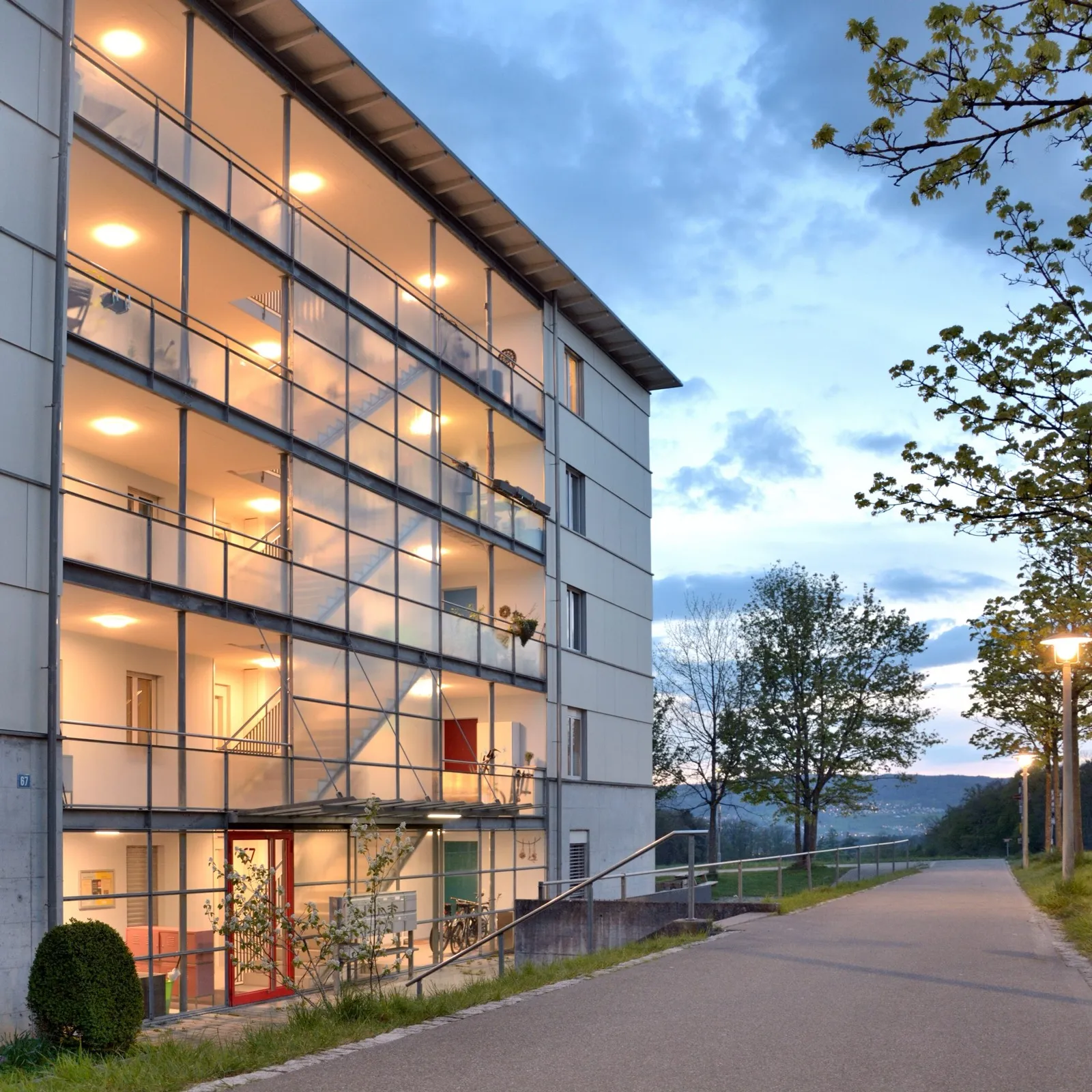 Rütihof-Gebäude mit beleuchtetem Weg bei Dämmerung, ASIG Zürich-Höngg